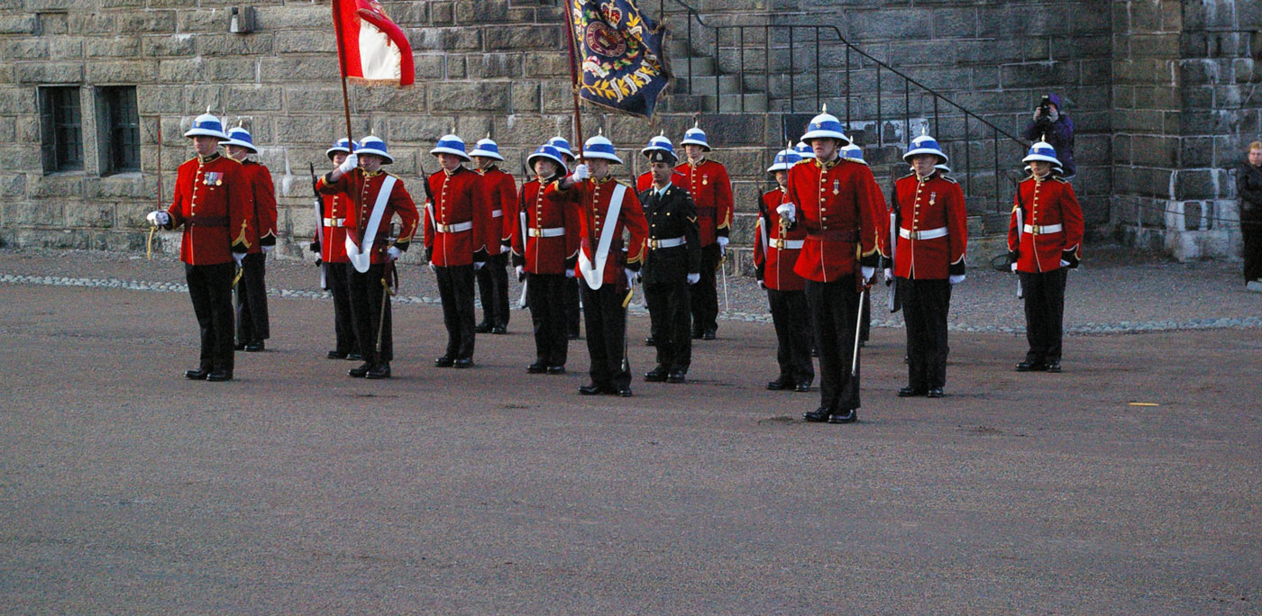 A group of ceremonial guards in red uniforms, white belts, and pith helmets stand in formation. Two guards hold flags in front of a stone building.