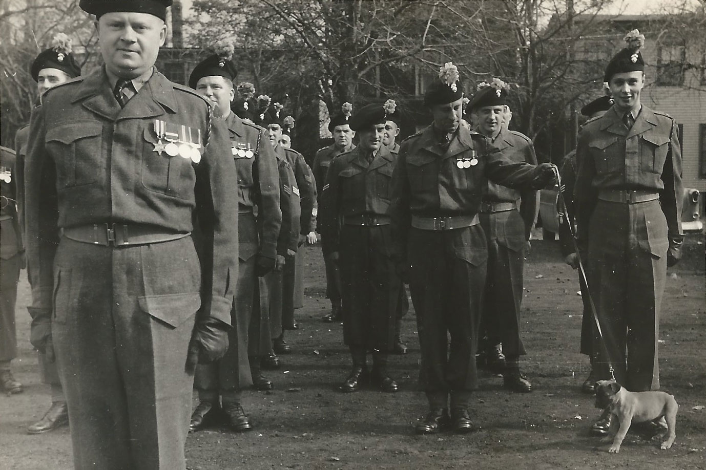 A group of uniformed soldiers stands in formation outdoors; one soldier holds a leash attached to a small dog.
