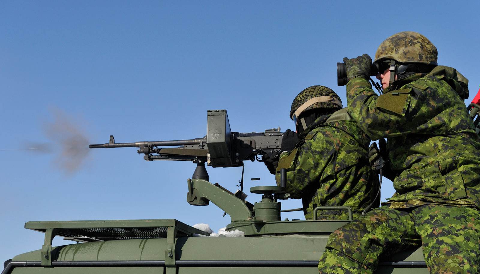 Two soldiers in camouflage uniforms are on a military vehicle, one firing a machine gun while the other uses binoculars.